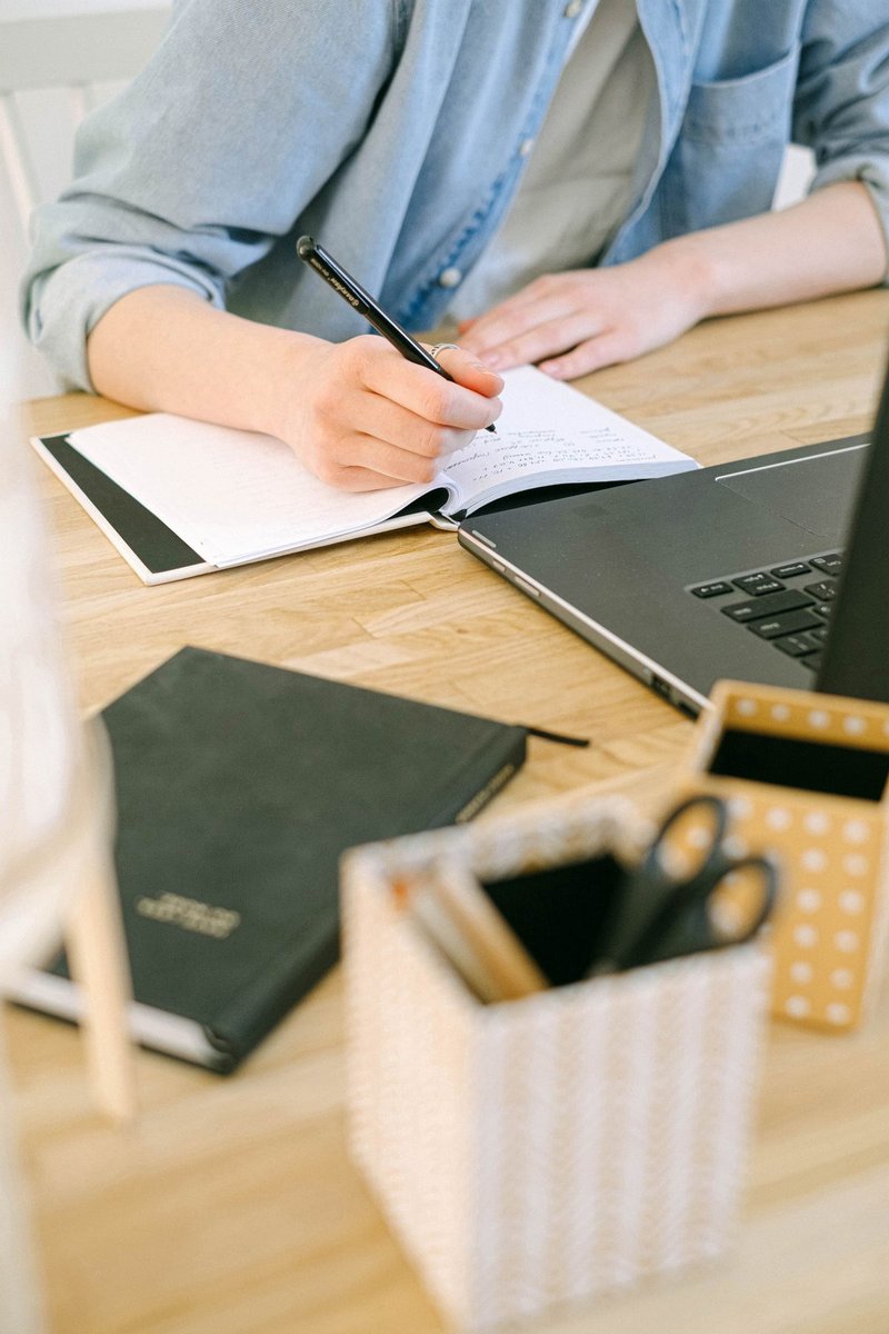 Person in a denim shirt writing in a small black notebook beside a laptop on a wooden desk